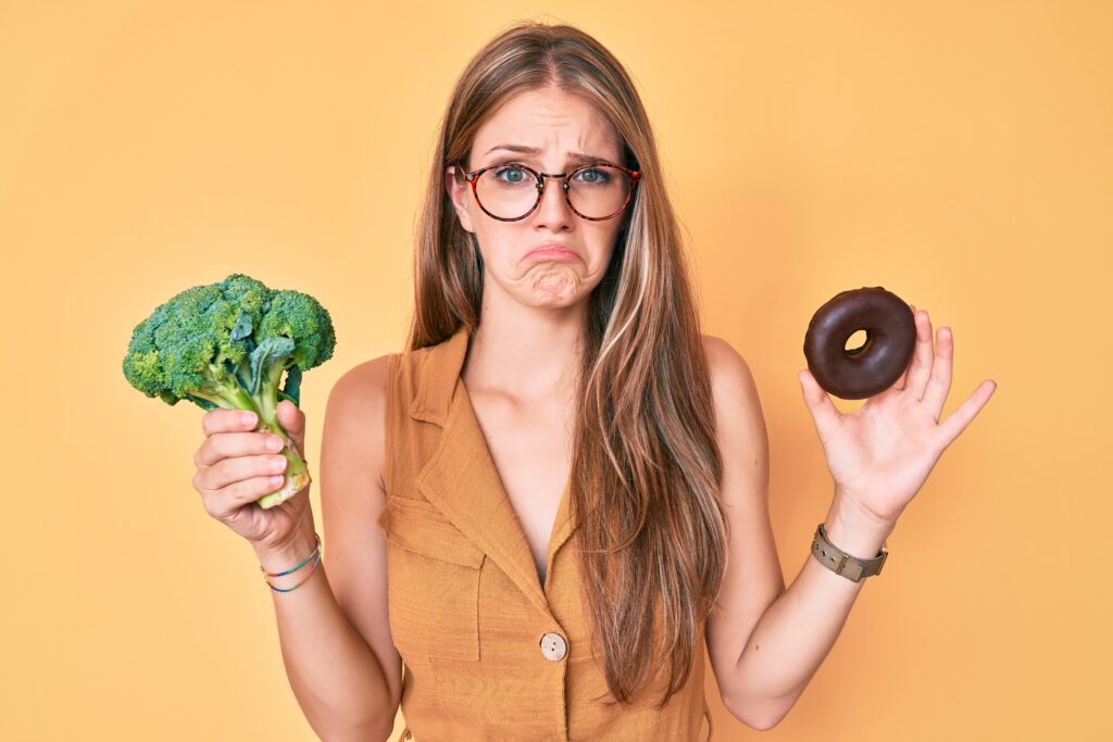 Non dimagrisci perché mangi troppo poco: un errore comune nella nutrizione sportiva young blonde girl holding broccoli and chocolate donut depressed and worry for distress, crying angry and afraid. sad expression.